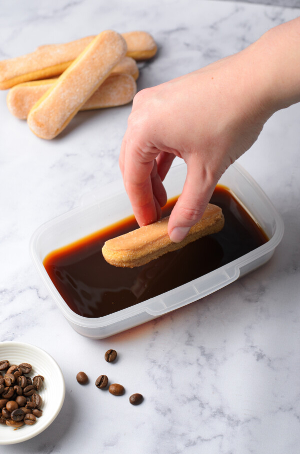 A woman's hand dipping a ladyfinger cookie into a small dish of coffee.