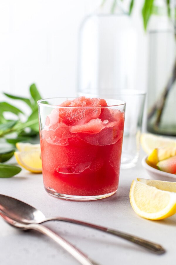 Watermelon frose in a cocktail glass, with fresh lemon wedges and mint leaves scattered over the table.