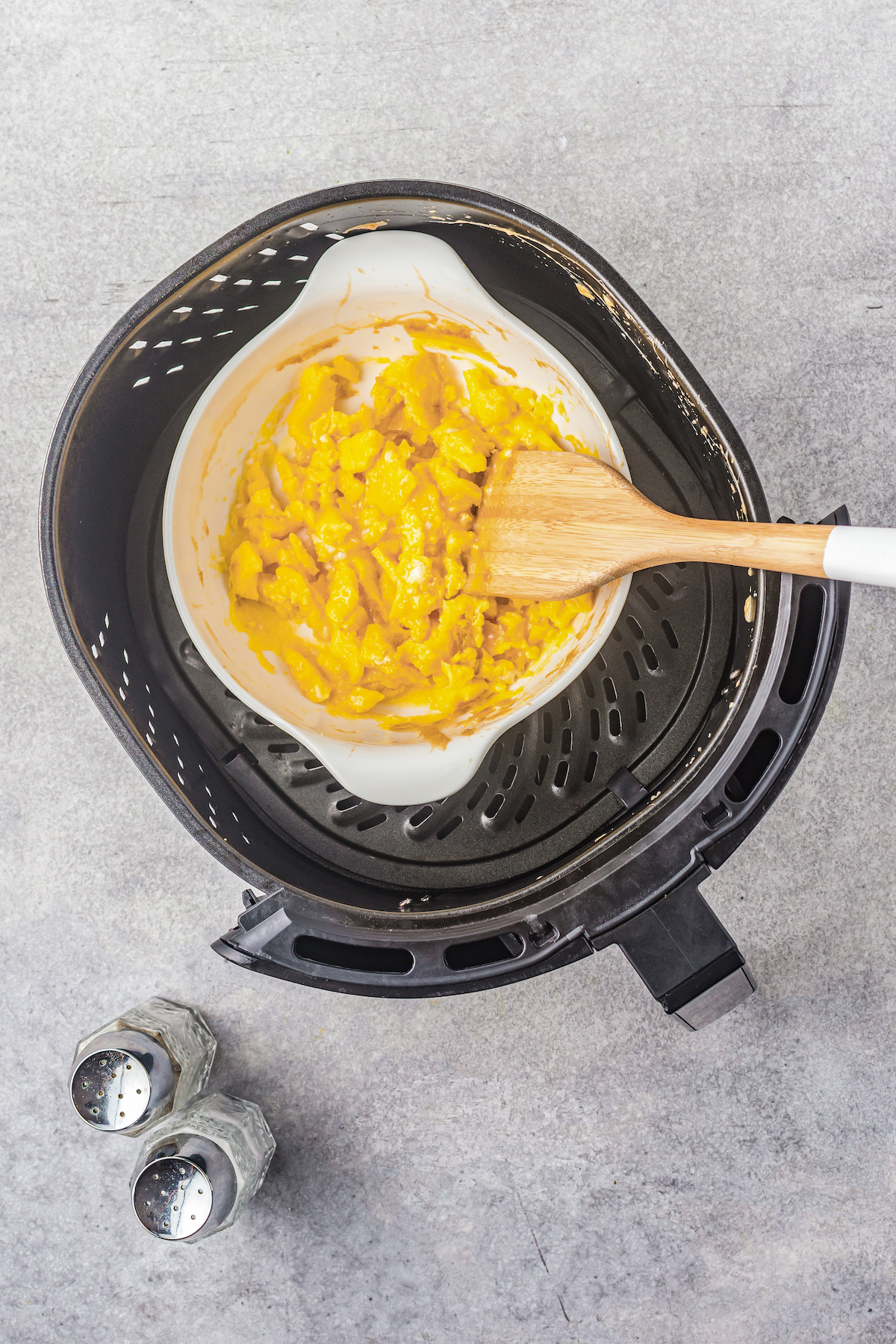 A wooden spoon is used to scramble eggs in a pot inside an air fryer.