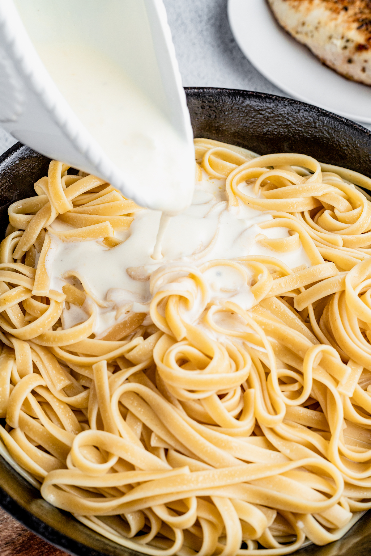 Creamy sauce being poured from a pitcher over a skillet full of cooked pasta.