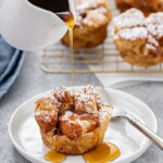 Syrup being poured from a small white pitcher onto a breakfast muffin. More muffins are on a cooling rack in the background.