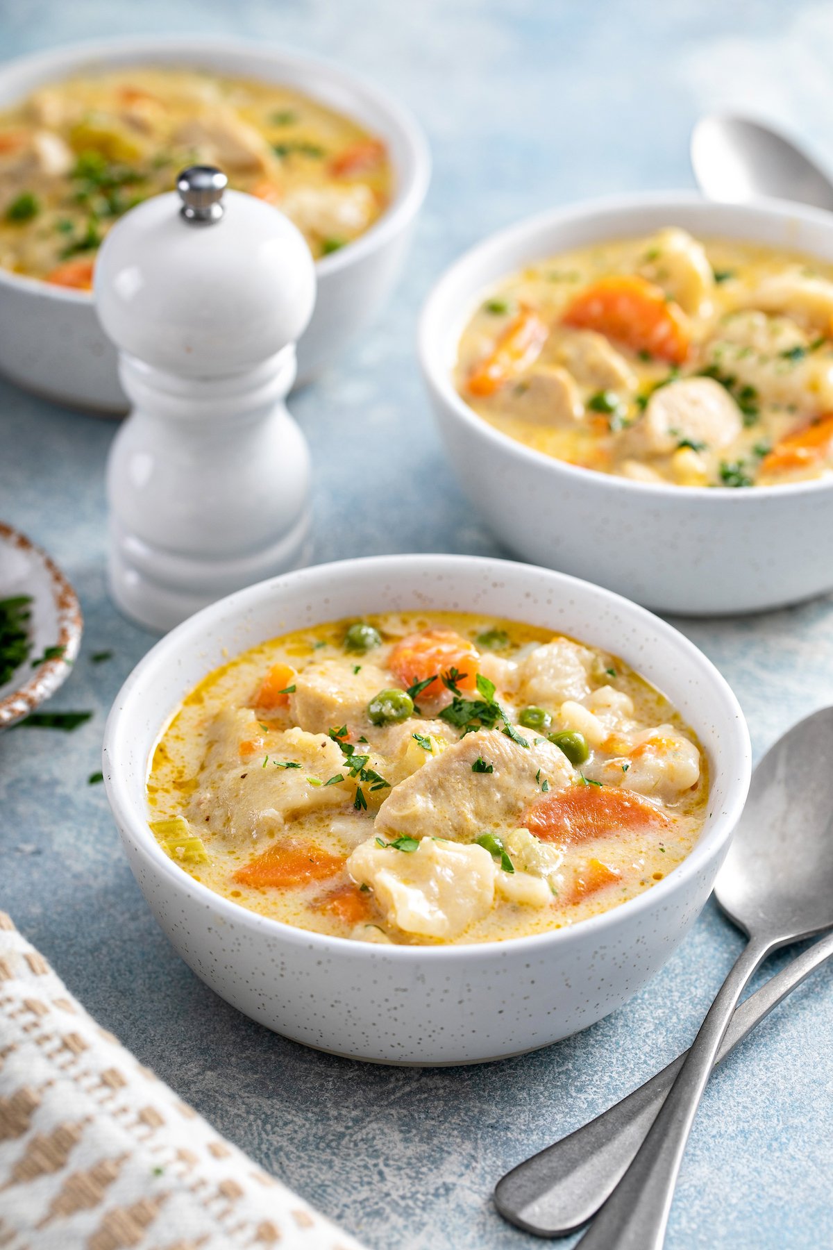 Three bowls of creamy chicken and veggies. Spoons and a white pepper grinder are on the table.