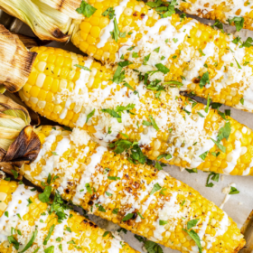 6 cobs of corn with Crema, cilantro, and seasonings on a sheet pan with parchment paper.