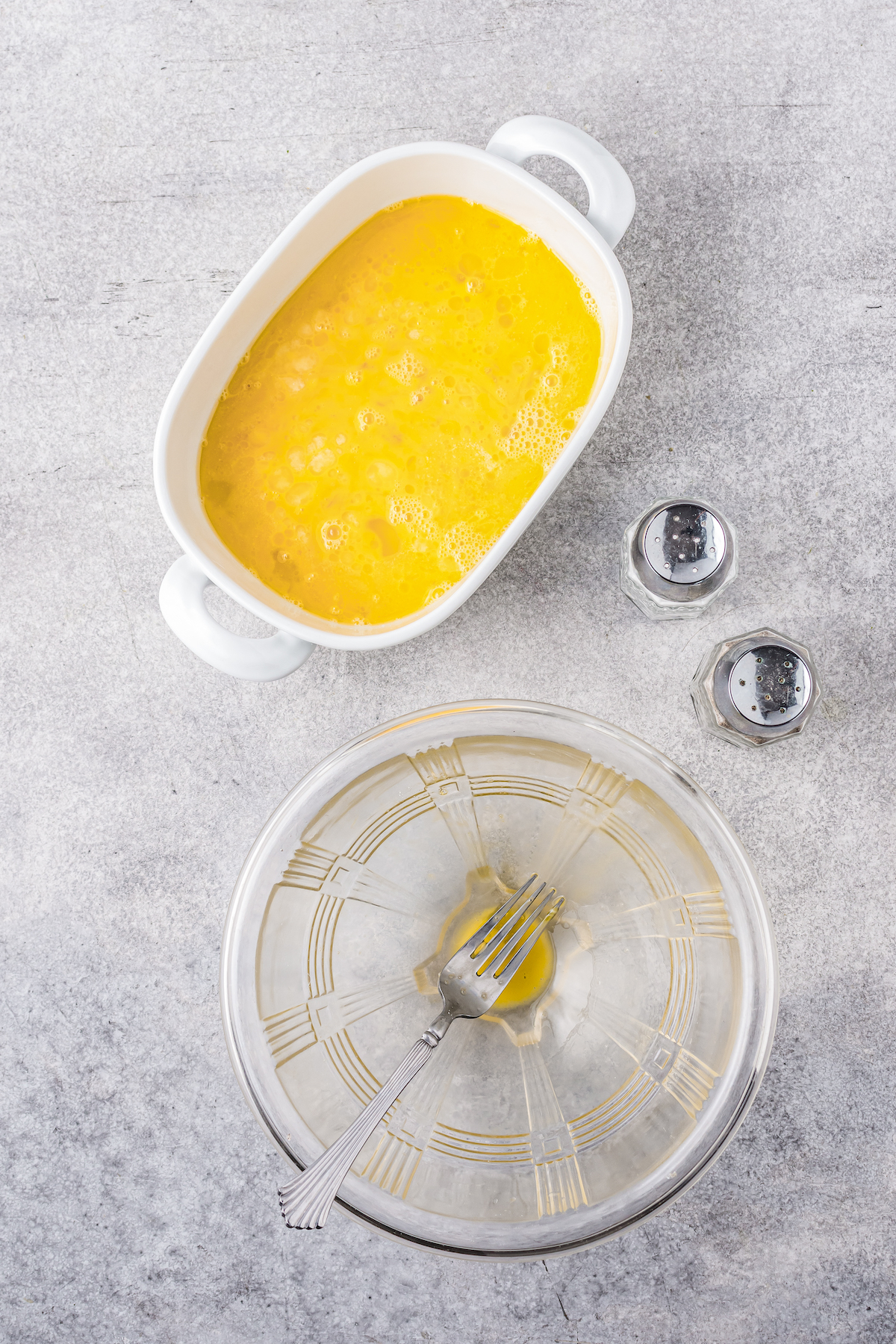 A white dish filled with whisked eggs next to a clear mixing bowl and salt and pepper holders.