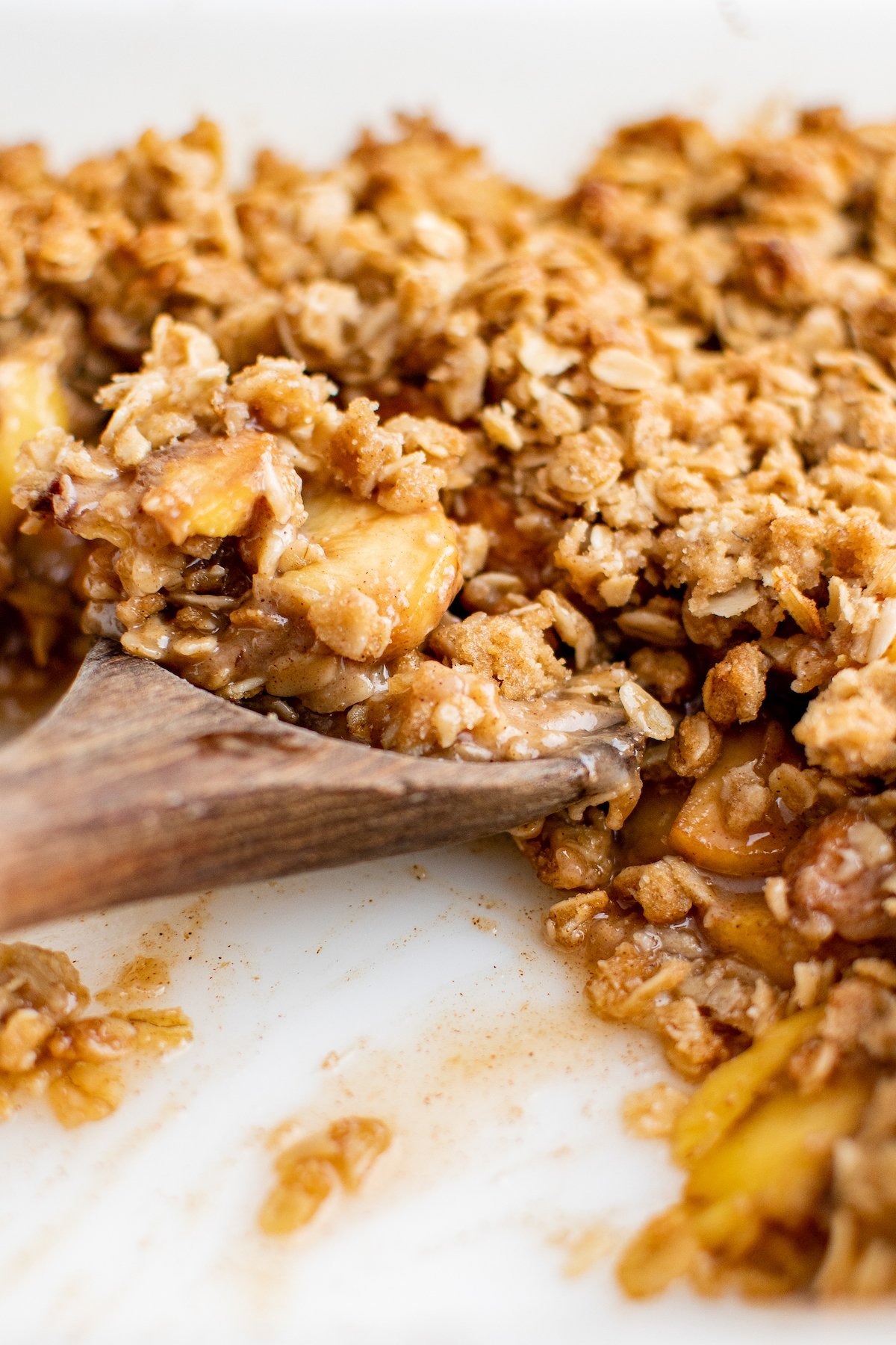 A wooden spoon digging into a baked peach dessert with oat topping.