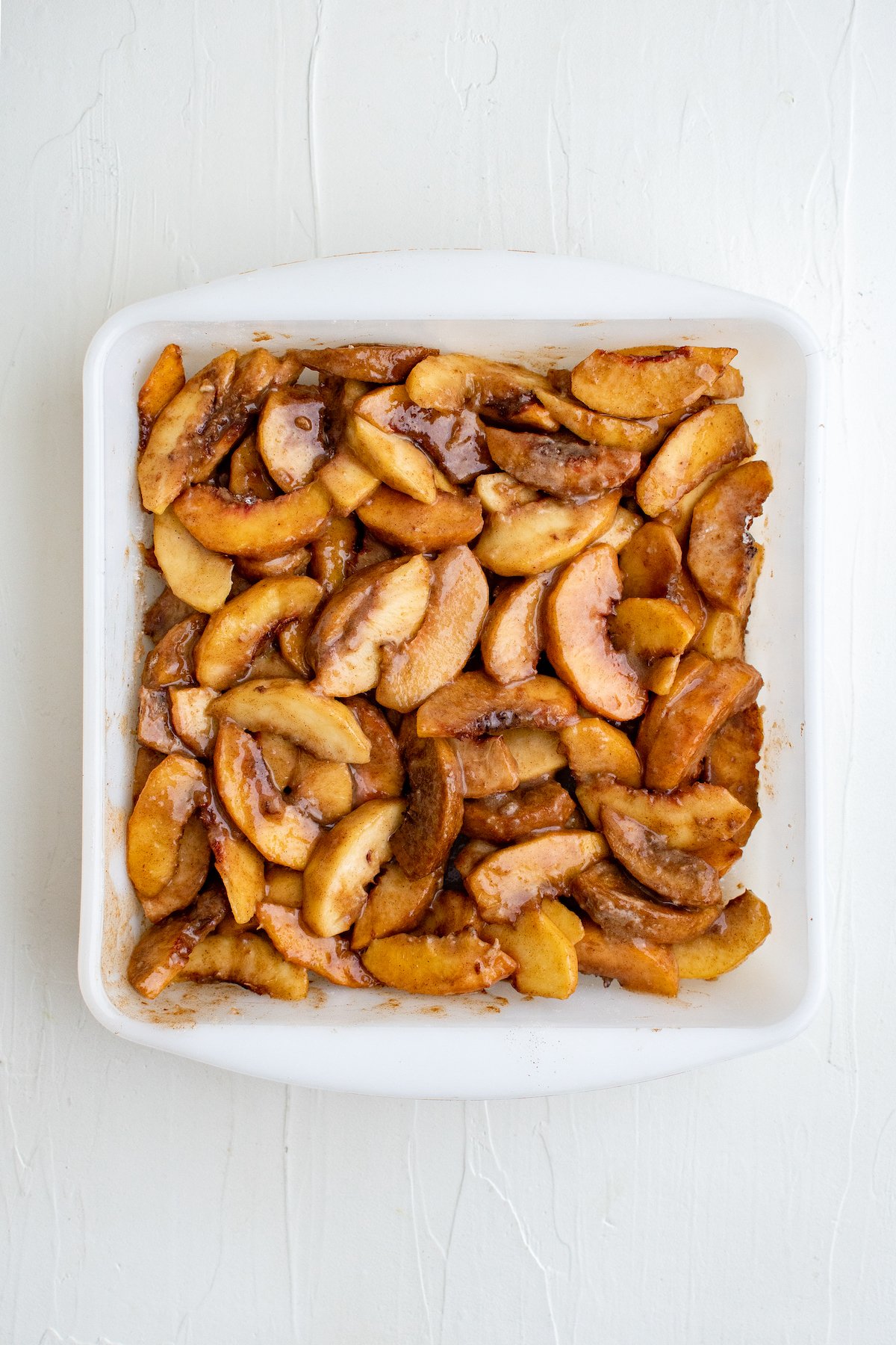 A square white baking dish with a layer of raw peaches in the bottom. The peaches are coated in a cinnamon-flour mixture.