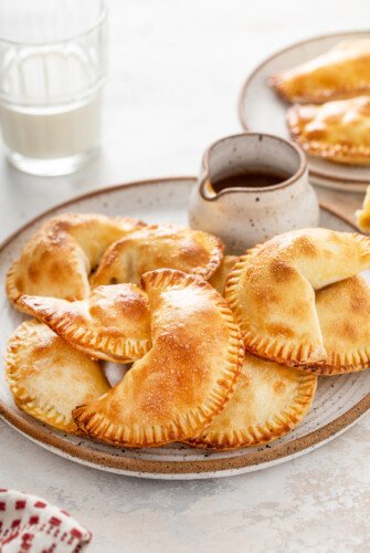 Golden-brown baked pineapple empanadas on a platter, with a small pitcher of caramel sauce.