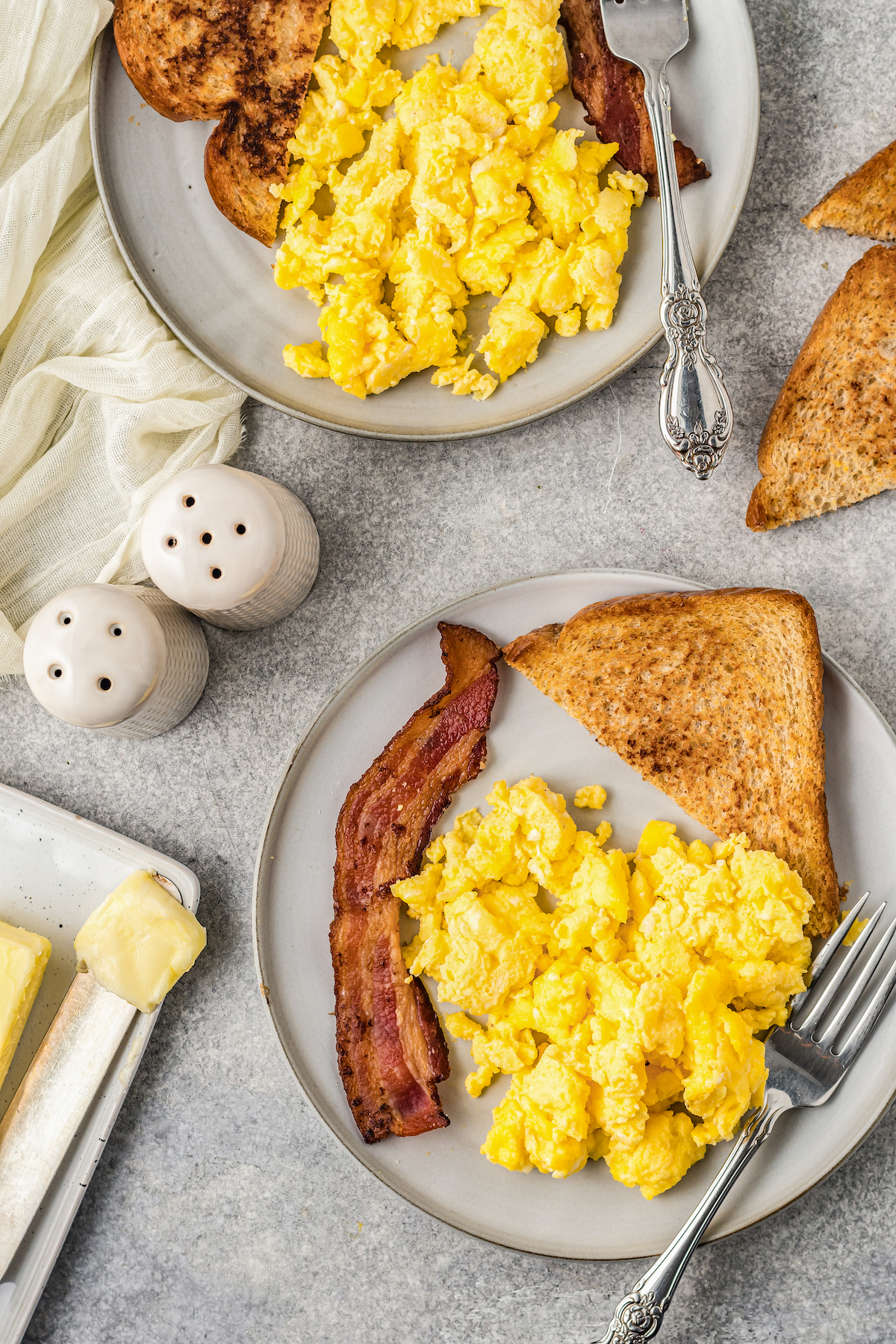 A breakfast spread of scrambled eggs on plates, next to toast and bacon.
