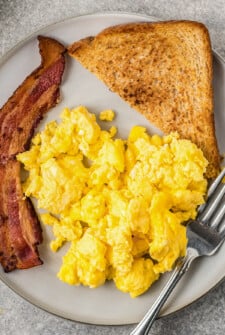 Top view of scrambled eggs on a plate next to toast and bacon.