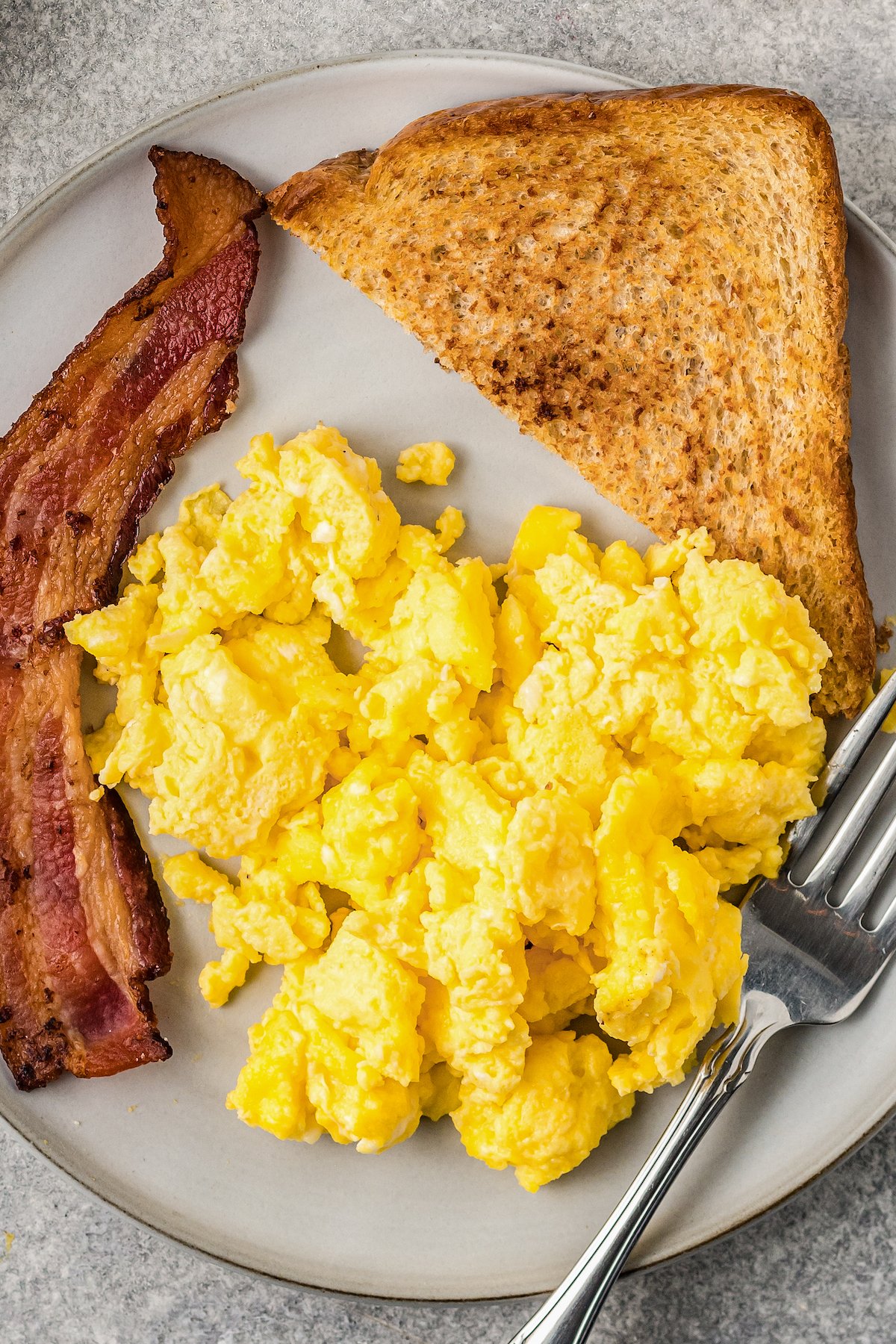 Top view of scrambled eggs on a plate next to toast and bacon.