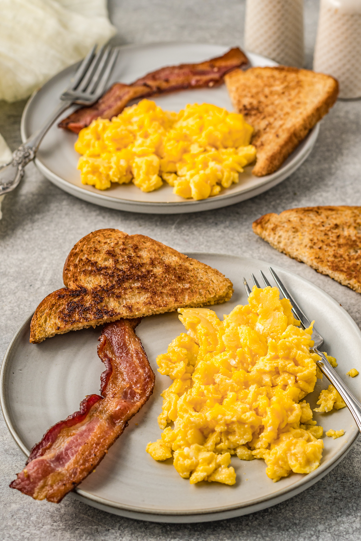 Two plates of stove top scrambled eggs with slices of toast and bacon.