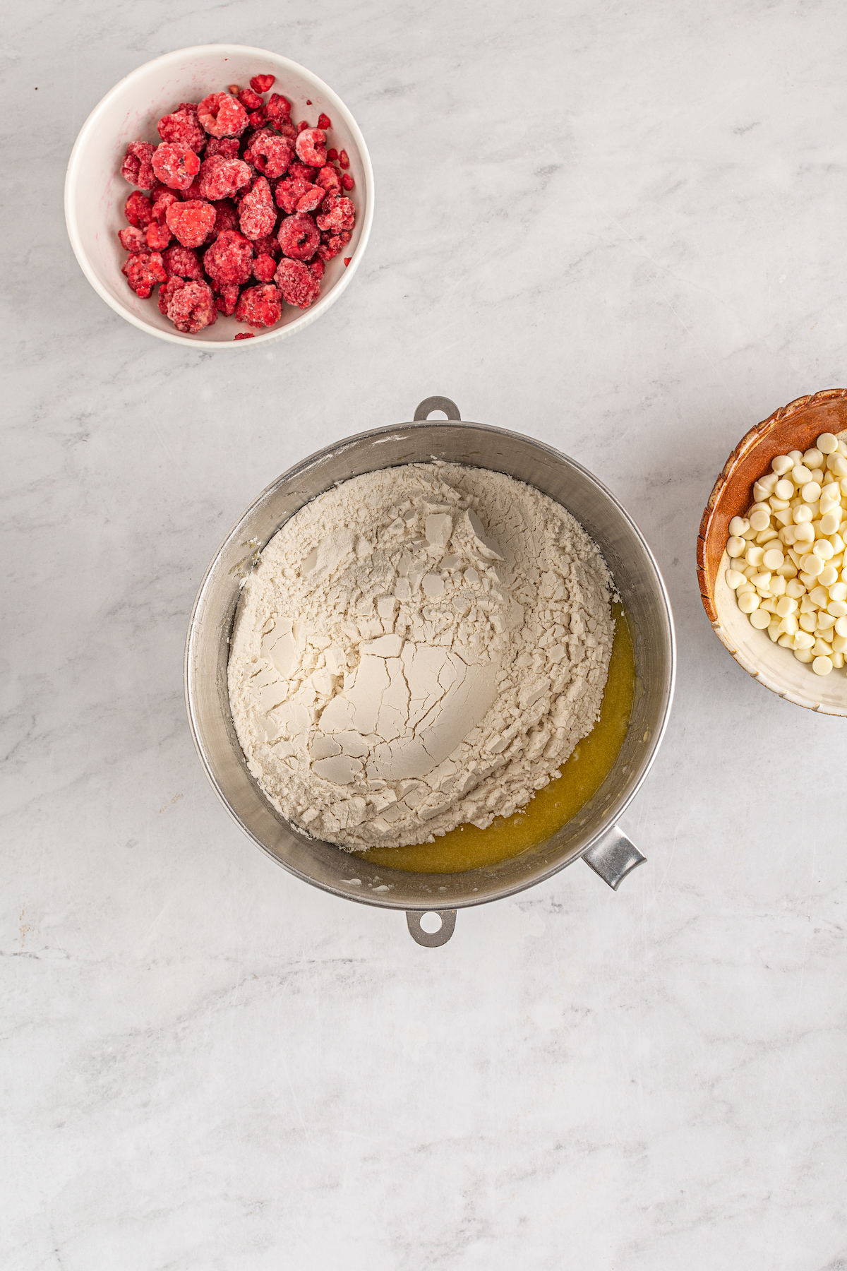 Dry ingredients on top of wet ingredients, in a metal mixing bowl. Raspberries and white chocolate chips are in bowls nearby on the work surface.