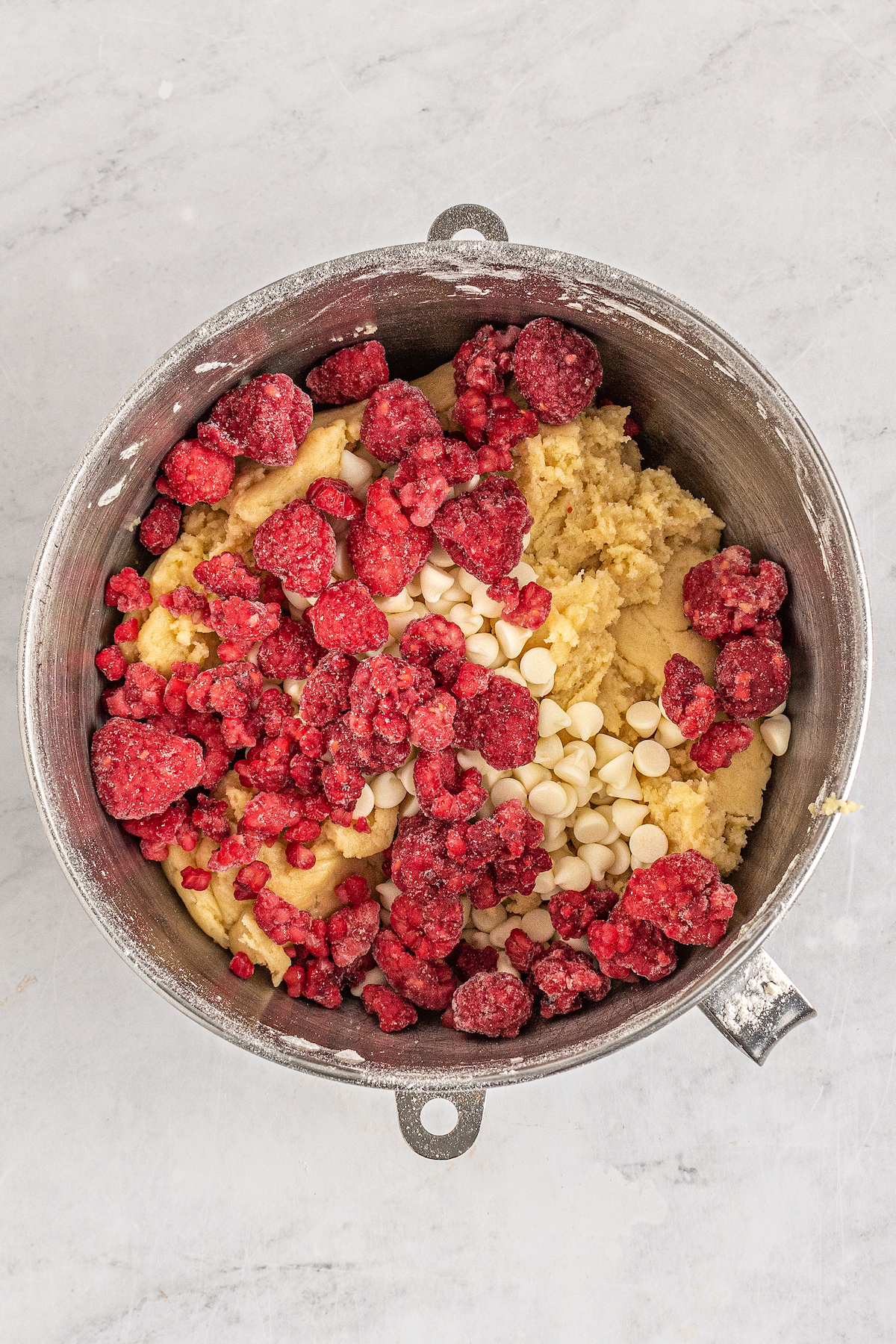 Cookie dough in a mixing bowl. Frozen raspberries and white chocolate chips have been dumped on top, but are not yet mixed in.
