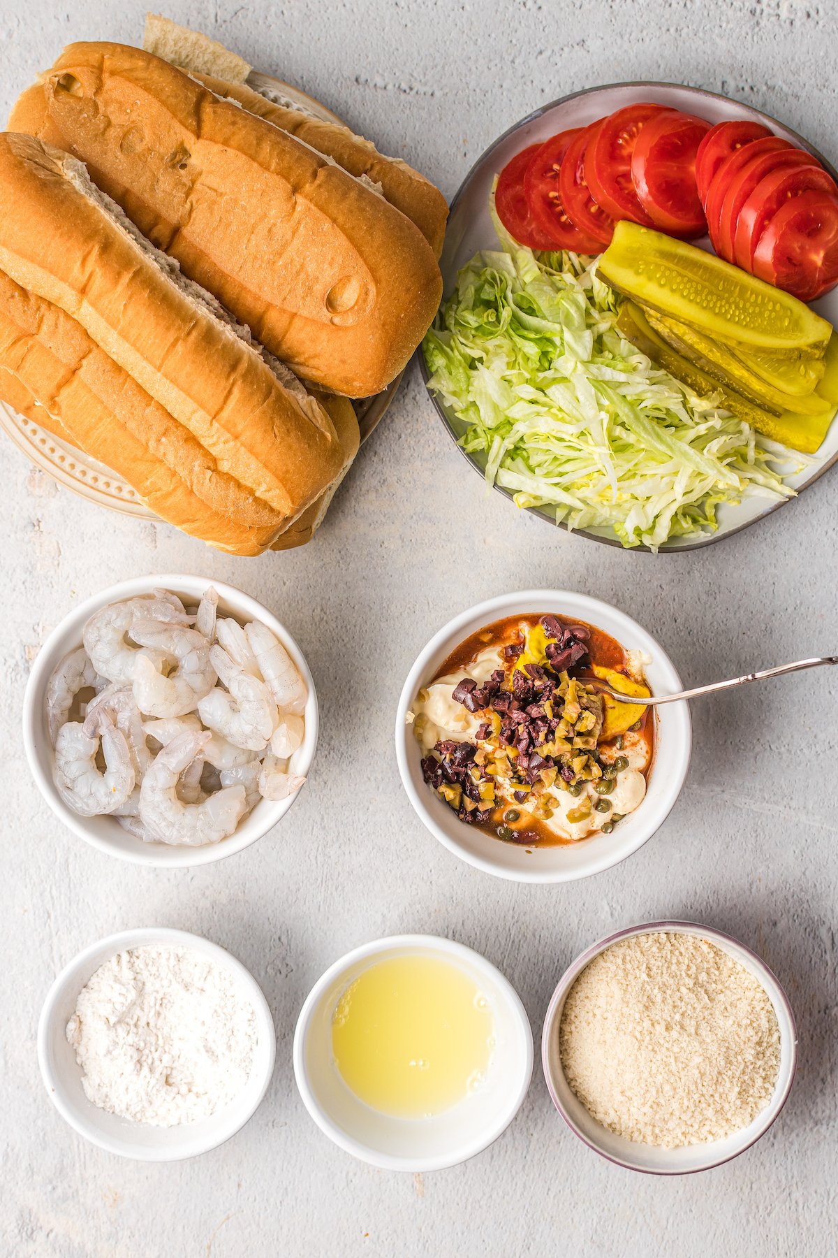 From top left: Sandwich buns, lettuce, tomato slices, pickle slices, raw shrimp, a bowl with remoulade ingredients and a spoon, and three bowls of flour, egg whites, and panko.