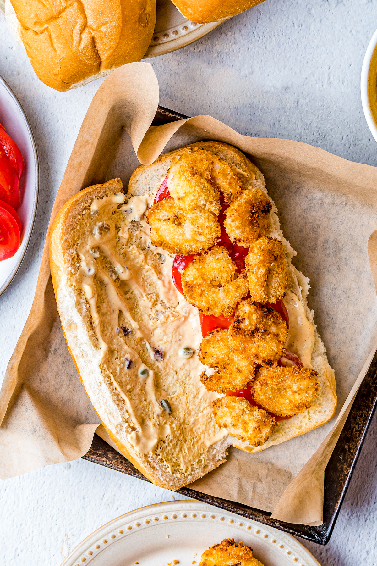 A sandwich bun topped with remoulade, sliced tomatoes, and breaded fried shrimp.
