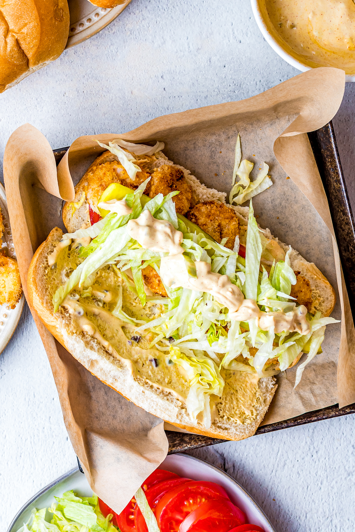 A sandwich bun sliced open and topped with sandwich spread, fried shrimp, shredded lettuce, and tomato.