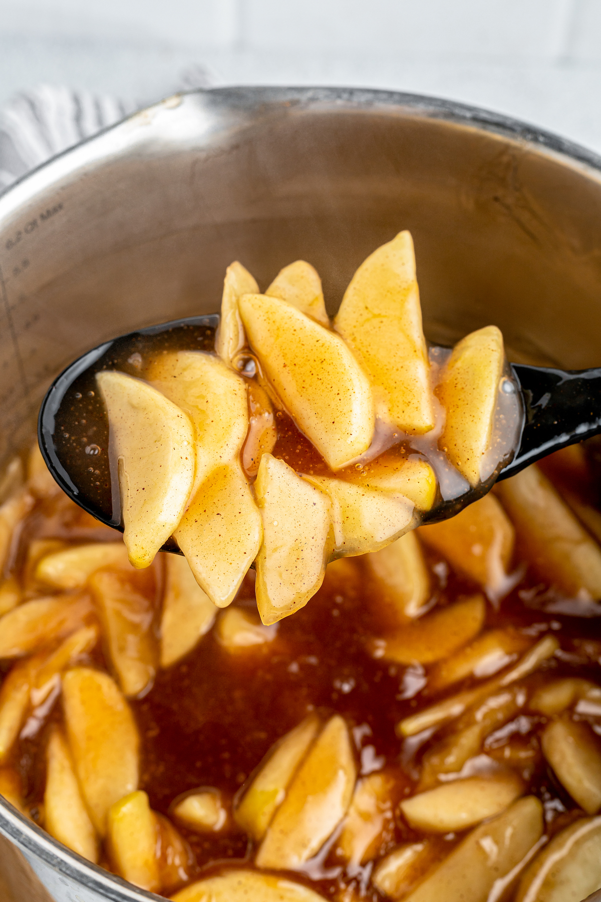 A spoon lifts cooked apple pie filling from a pot of apples simmering in sauce.