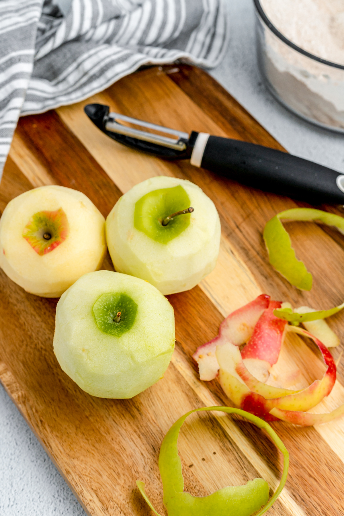Three peeled apples on a wooden cutting board, next to apple skins and a peeler.