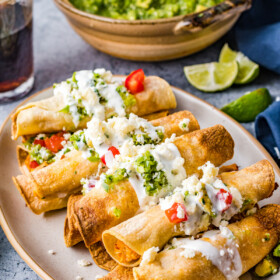 Chicken taquitos arranged on a platter and garnished with guacamole, crema, and more.