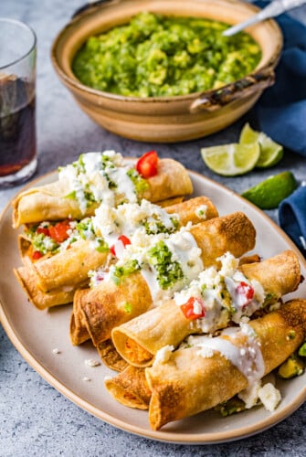 Chicken taquitos arranged on a platter and garnished with guacamole, crema, and more.