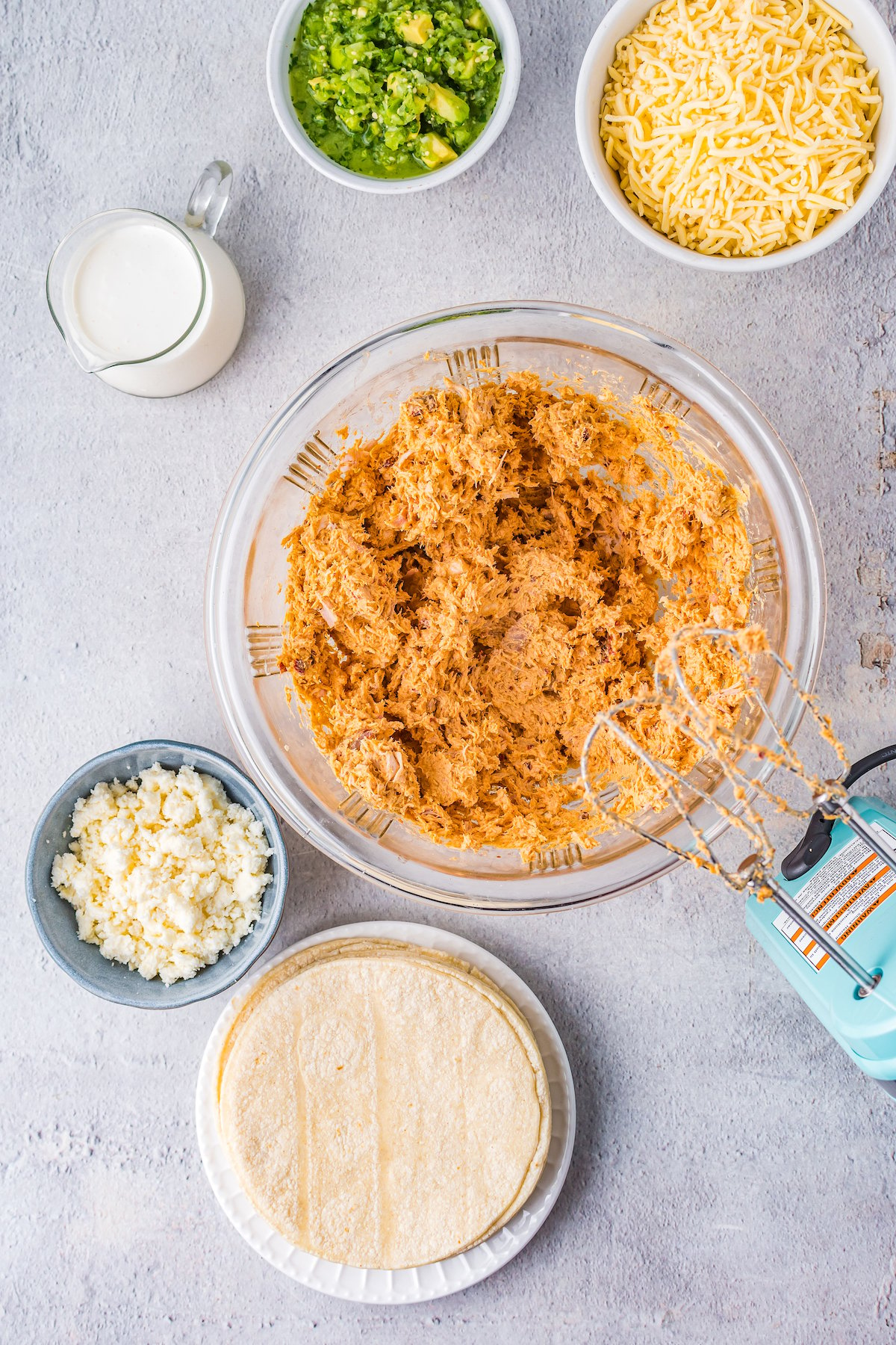 Dishes of crema, guacamole, shredded cheese, cotija cheese crumbles, and tortillas are arranged around a large mixing bowl of creamy chicken filling.