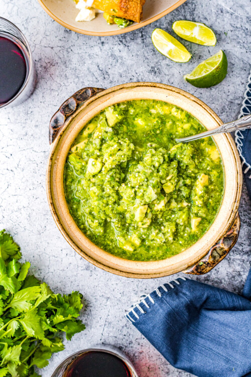 A bowl of green salsa on a countertop. Dishes of ingredients, a bunch of cilantro, and a cloth napkin are arranged around the bowl.