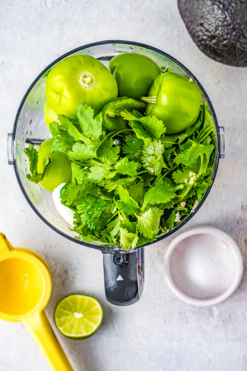 Overhead shot of a food processor with tomatillos, cilantro, and other ingredients inside.