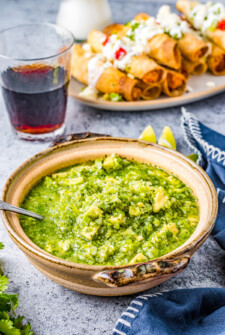 Salsa verde and a plate of taquitos on a table with a glass of red wine.