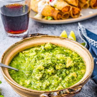 Salsa verde and a plate of taquitos on a table with a glass of red wine.