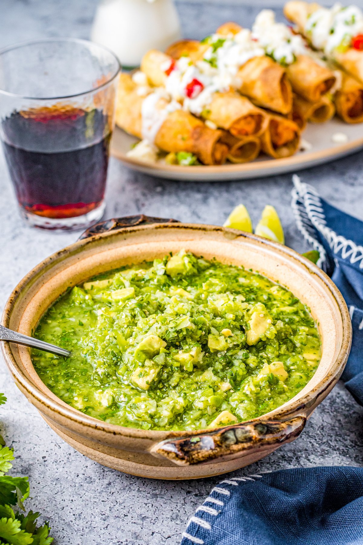 Salsa verde and a plate of taquitos on a table with a glass of red wine.