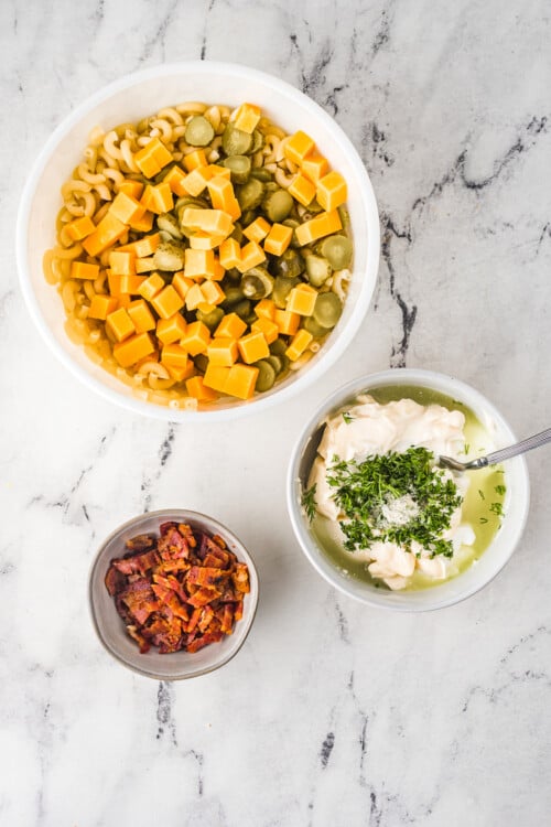 Cheese and pickles in a bowl of elbow pasta, next to a dish of dressing ingredients and another dish of bacon bits.