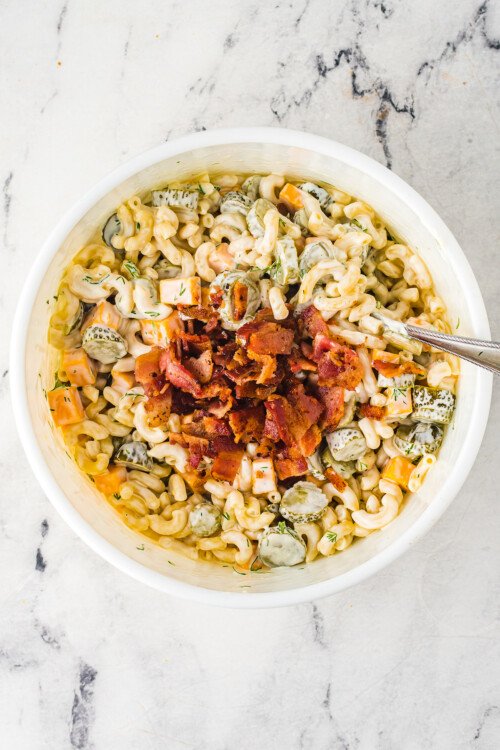 Dill pickle pasta salad in a serving bowl, with bacon being stirred in.