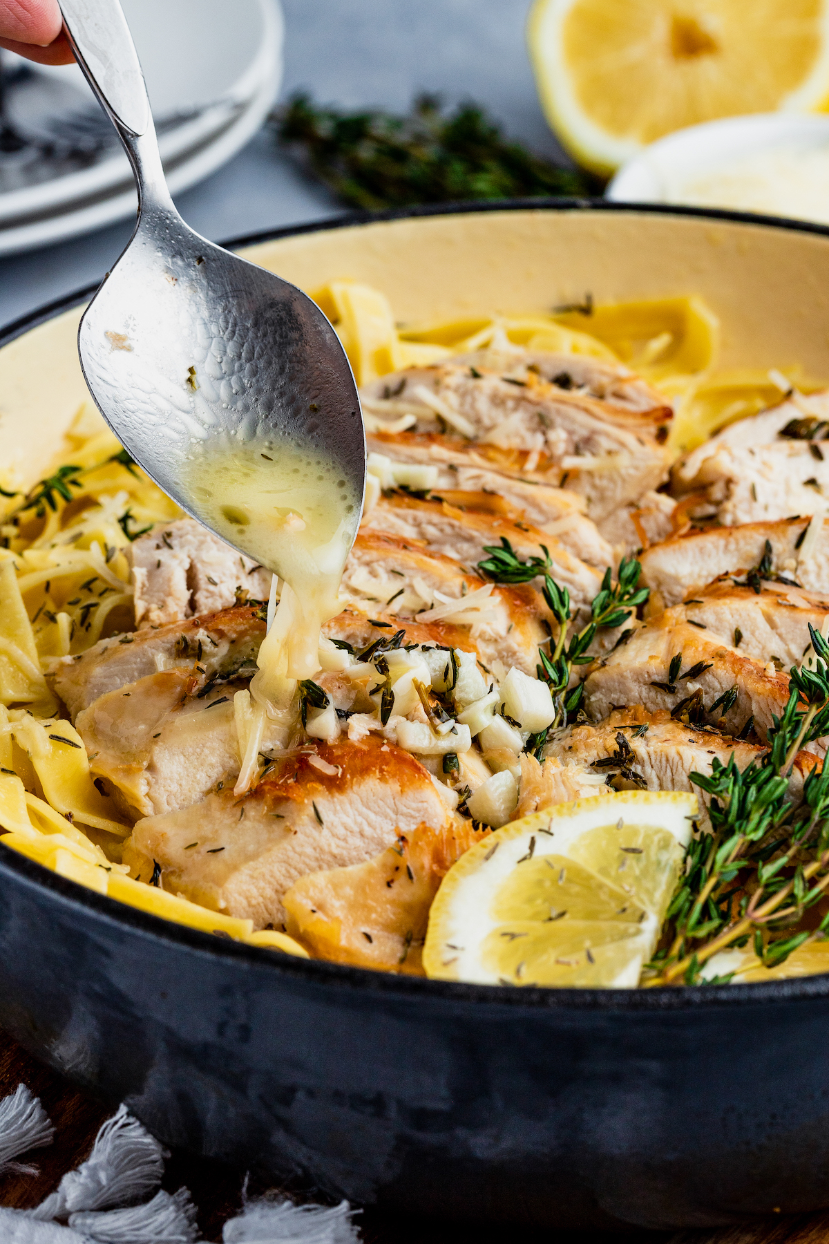 A spoon drips sauce overtop a bowl of lemon chicken pasta garnished with a lemon slice.