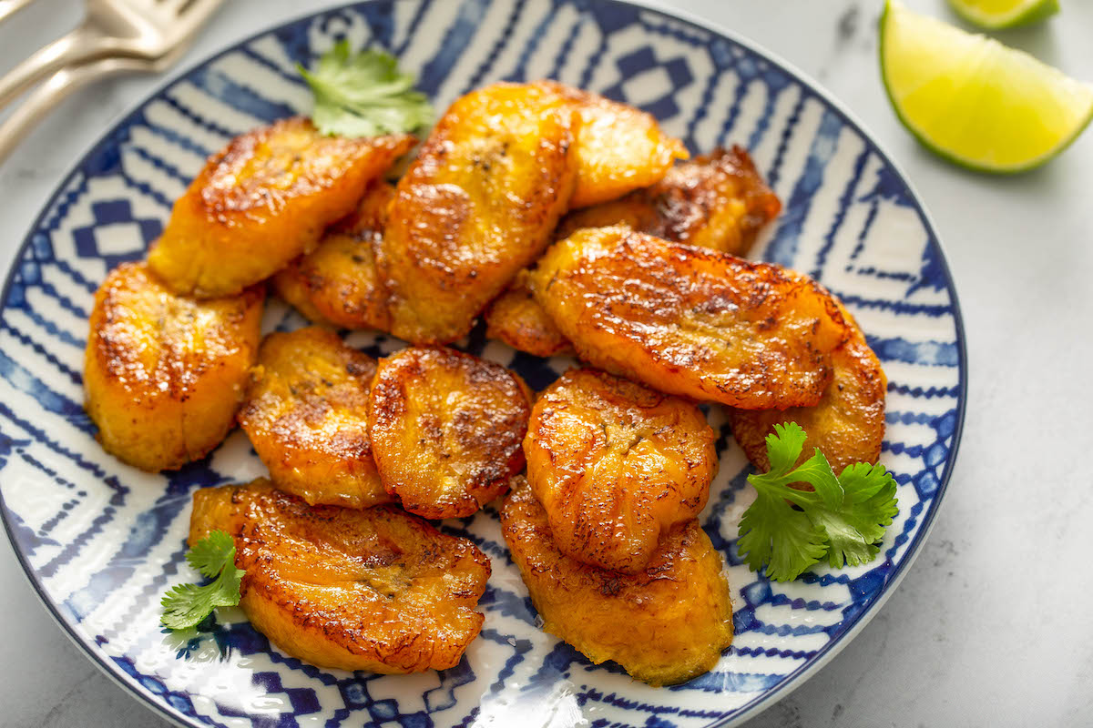 Overhead image of sweet plantains in a blue and white bowl with lime wedges and cilantro.