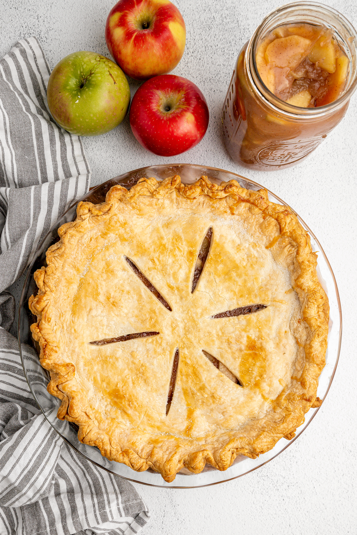 Top view of a baked apple pie next to a jar of apple pie filling and three apples.