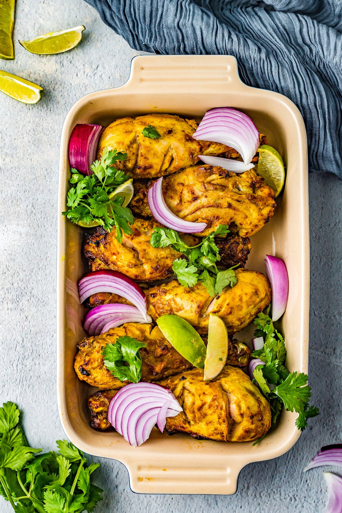 Baked tandoori chicken in a serving dish, garnished with herbs and red onion wedges.