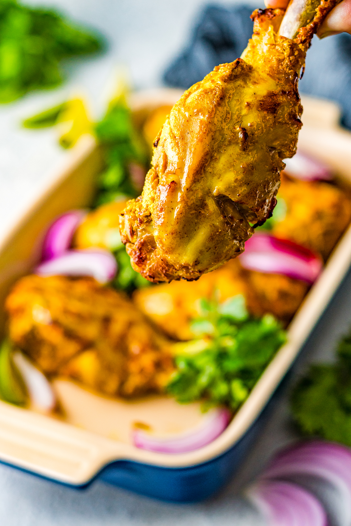 A piece of tandoori chicken is held near the camera to show texture. In the background is a serving dish of more chicken.