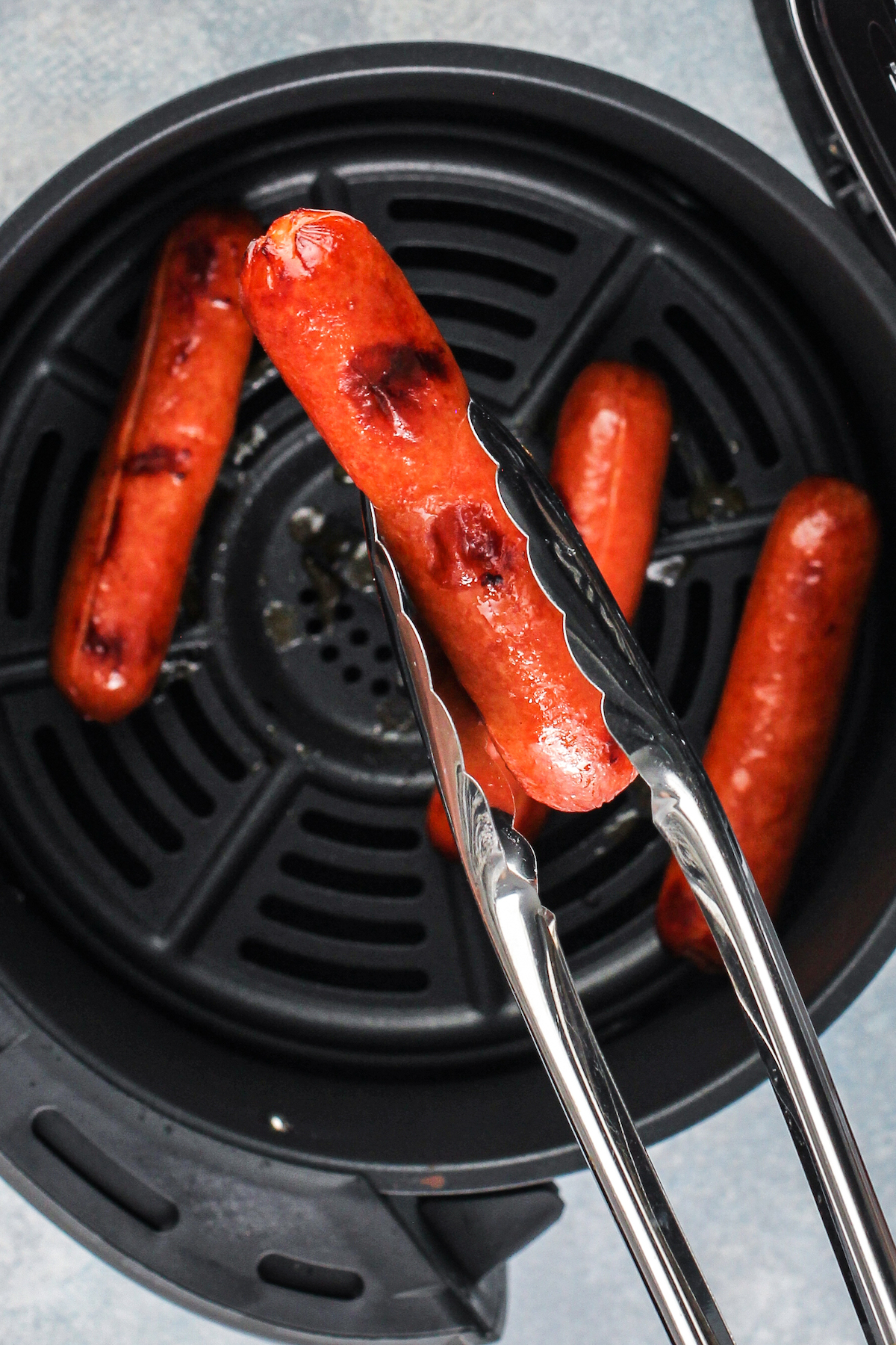 Metal tongs hold a roasted hot dog over the other hot dogs in the basket of an air fryer.