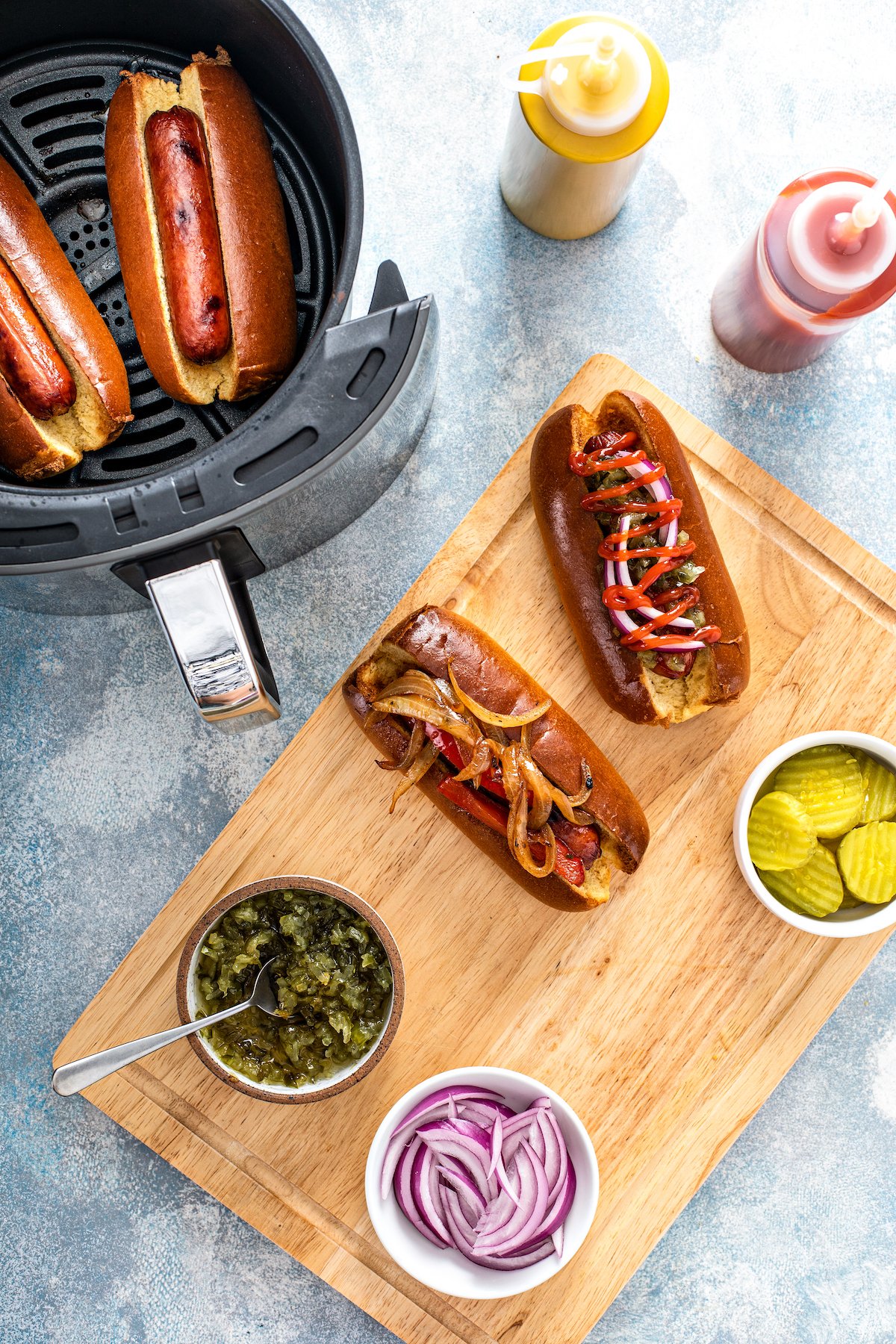 Top view of two hot dogs in buns on a wooden board next to various hot dog toppings, with two more hot dogs in the air fryer nearby.