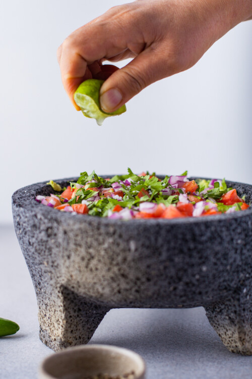 A lime being squeezed over a Mexican mortar and pestle full of guacamole ingredients.