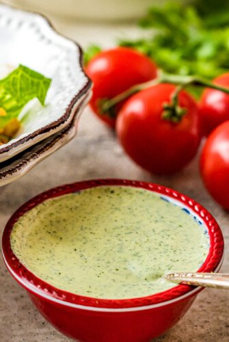 A small red bowl filled with creamy green salad dressing, next to a bowl of salad ingredients and a few fresh tomatoes.