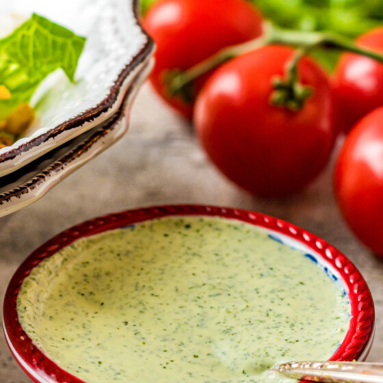 A small red bowl filled with creamy green salad dressing, next to a bowl of salad ingredients and a few fresh tomatoes.