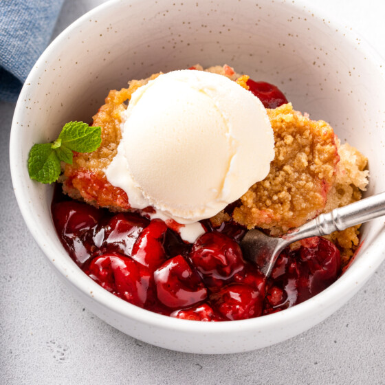 An up close image of cherry cobbler in a bowl with ice cream and a spoon scooping up the cherry filling.