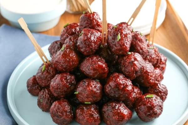 Easy 3-ingredient Grape Jelly Meatballs made in the crockpot or on the stovetop. Sweet, savory, and gone in minutes at every party! A stack of glazed grape jelly meatballs with toothpicks, garnished with herbs, is arranged on a light blue plate.