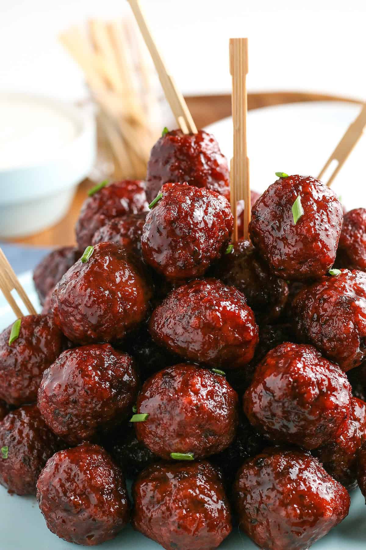 Easy 3-ingredient Grape Jelly Meatballs made in the crockpot or on the stovetop. Sweet, savory, and gone in minutes at every party! A close-up of glazed grape jelly meatballs stacked on a place with a few meatballs on top pierced with a wooden skewer.