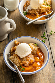 A bowl of crockpot peach cobbler topped with vanilla ice cream on a wooden table, with a second bowl in the background.
