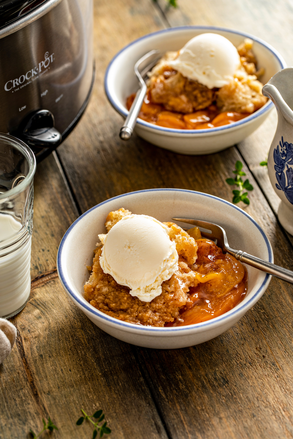 Two bowls of crockpot peach cobbler servings topped with vanilla ice cream, on a wooden tabletop.