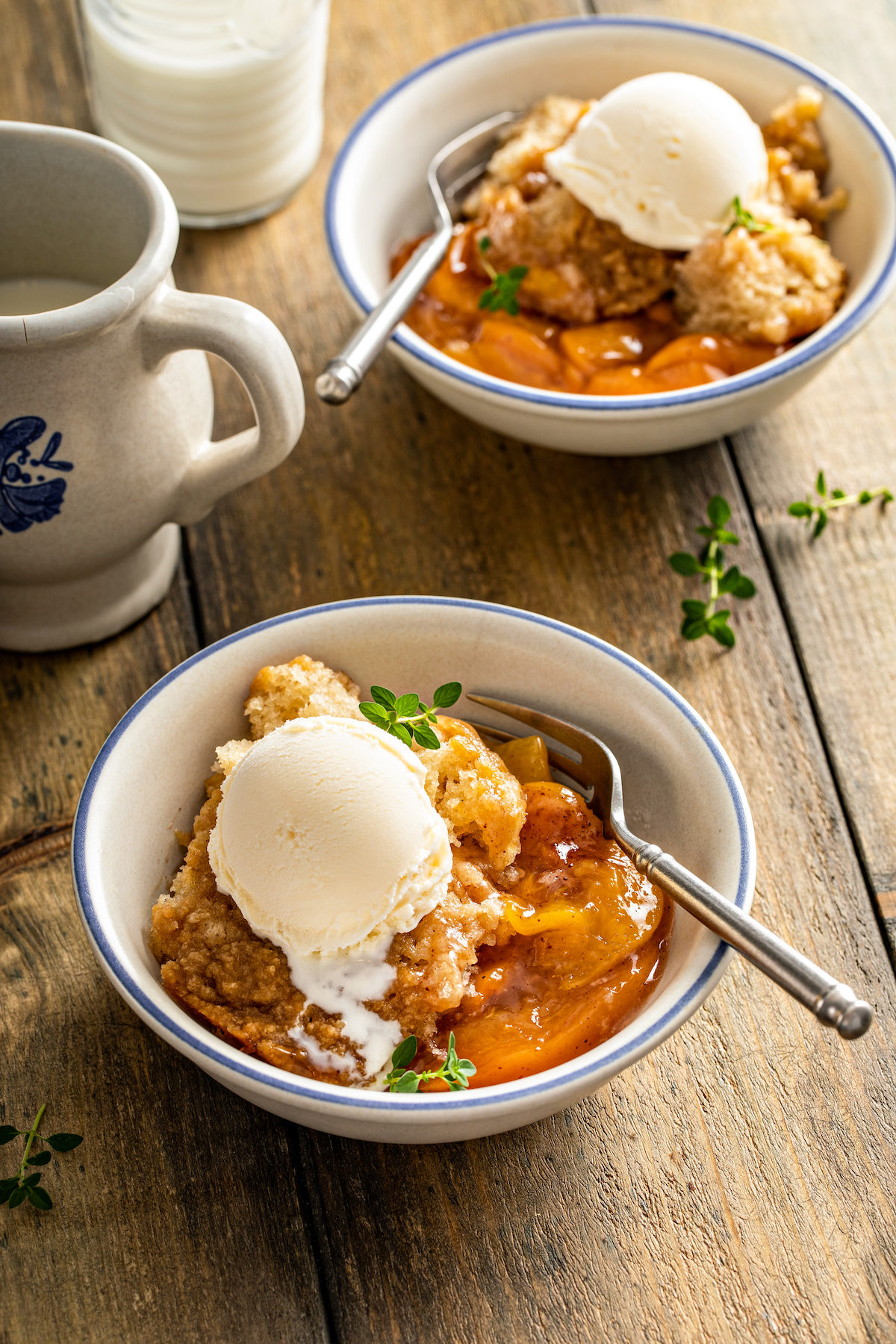 Two bowls of crockpot peach cobbler servings topped with vanilla ice cream, on a wooden tabletop.