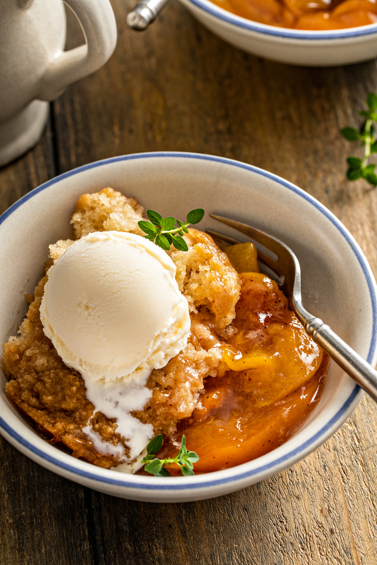 Close up of a bowl of crockpot peach cobbler topped with vanilla ice cream and garnished with thyme sprigs, with a spoon.