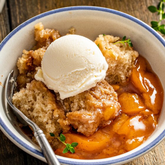 Close up of a bowl of crockpot peach cobbler topped with vanilla ice cream and garnished with thyme sprigs, with a spoon.